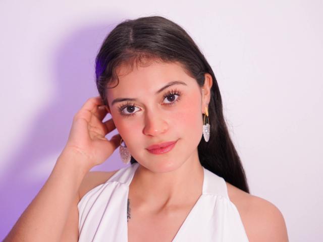LizzaRose has long dark hair, wearing a white halter top, pink blush, and diamond earrings, touching her head with a gentle smile, pink lips, and brown eyes.