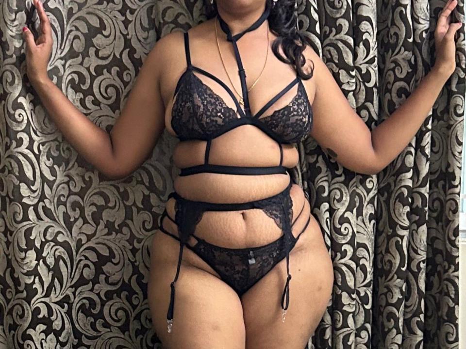 Shruthi, avec un corps aux formes généreuses, portant de la lingerie noire en dentelle avec des bretelles fines, montrant ses seins et ses parties intimes, une ceinture à jarretières et un soutien-gorge à bretelles fines, debout devant un rideau à motifs, les mains levées..
