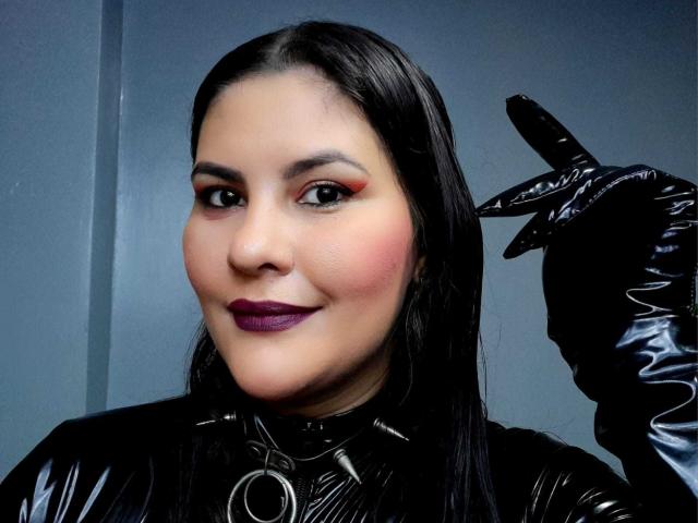 Sharlok, met zwarte latex handschoenen, rode en oranje oogschaduw, paars lippenstift en een zwart, glanzend latex outfit, maakt een gebaar met haar vinger en glimlacht. Haar achterkant is niet zichtbaar..