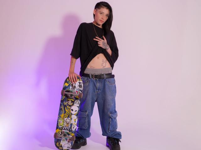MorganWallen with black shirt, lifted to show belly tattoos, holding colorful skateboard, wearing loose blue jeans, black boots, and a necklace, standing against white wall.