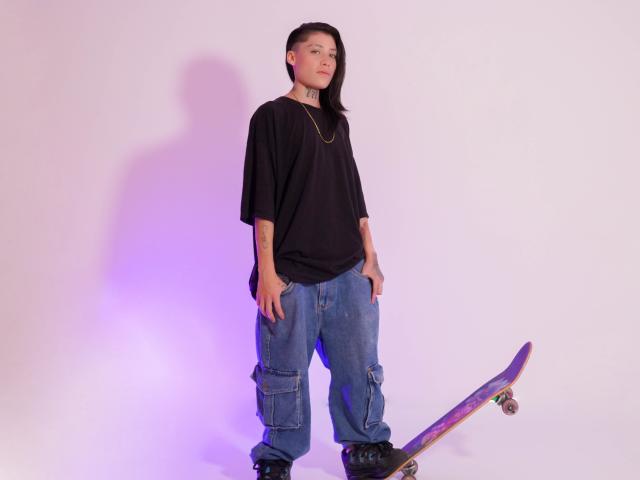 Morgan Wallen, avec des tatouages, portant une chemise noire, un jean bleu ample, des bottes noires, debout avec un skateboard, une ombre sur un mur blanc, une lumière violette sur la gauche..
