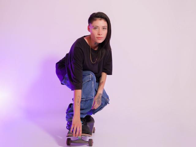 MorganWallen is crouched on a skateboard, wearing a black shirt, blue jeans, and a necklace, with tattoos on their arms, and has purple-tinted hair, looking at the camera.