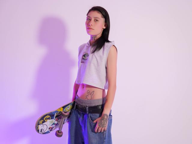 Morgan Wallen met lang zwart haar, een grijze crop top zonder mouwen, blauwe jeans, die een skateboard vasthoudt met een kleurrijke grip, tatoeages op de buik en hand, een paarse schaduw op een witte muur..
