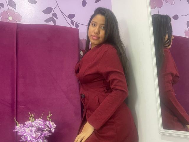 SaraMonrroe with long black hair, wearing a red coat, standing next to a purple velvet headboard, reflection in mirror, purple flowers in bottom left.