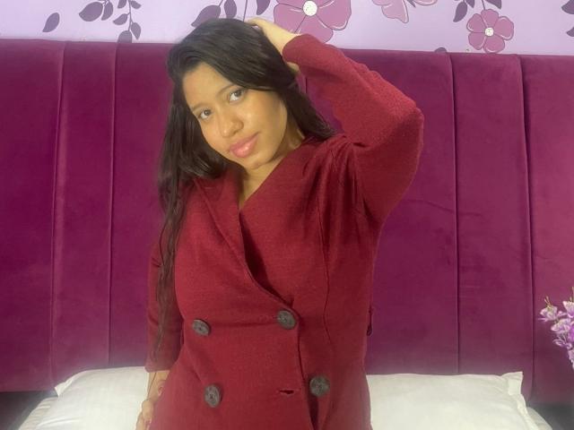 SaraMonrroe with long black hair, wearing a red button-up coat, standing in front of a purple velvet headboard, hand in hair, looking at camera, slight smile.