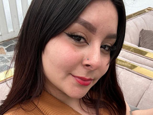 EmaJames has dark brown hair, a small nose piercing, red lipstick, and black eyeliner, smiling with a slightly upturned nose. She's wearing a brown top, looking cute.