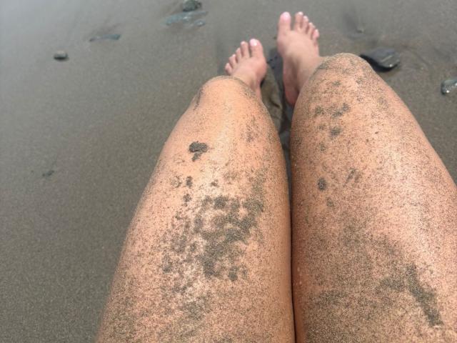 CarlaMuille's sand-covered legs with small patches of dirt, feet with pink toenails, and slightly parted toes, lying on a wet, sandy beach.