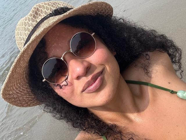 CarlaMuille with curly black hair, wearing a straw hat, round sunglasses, and a green necklace, has a smile and a small scar under her right eye, at the beach.