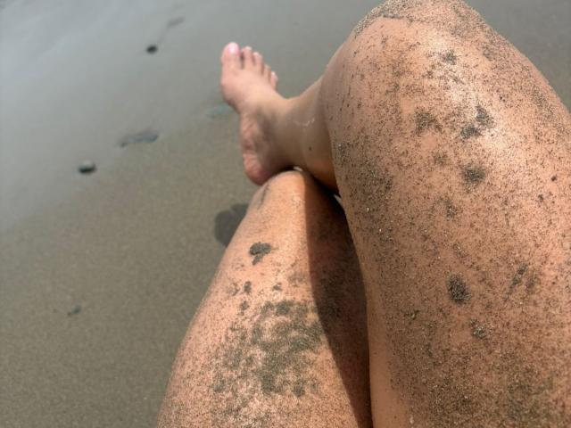 CarlaMuille's sandy-covered thighs and feet, showing small black sand patches, with her toes pointed slightly upwards. Her skin is tan and the sand is gritty.