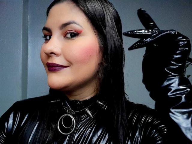 Sharlok with black latex gloves, shiny black latex top, and purple lipstick, showing a hand gesture, has pink cheeks, dark eyes, and straight black hair.