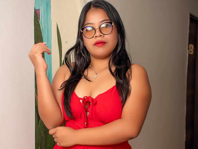 SelenaVaughn, met zwart haar, een rode jurk en een bril, heeft haar hand in de buurt van haar gezicht, waarbij haar borsten en een kleine ketting met een kruis zichtbaar zijn. Ze staat in een kamer..