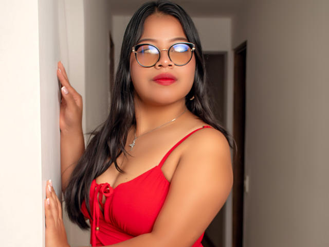 SelenaVaughn con un vestido rojo, lápiz labial rojo y gafas, de pie en un pasillo, tocando la pared con la mano derecha, con senos medianos, cabello negro largo, expresión segura, collar con un pequeño dije..