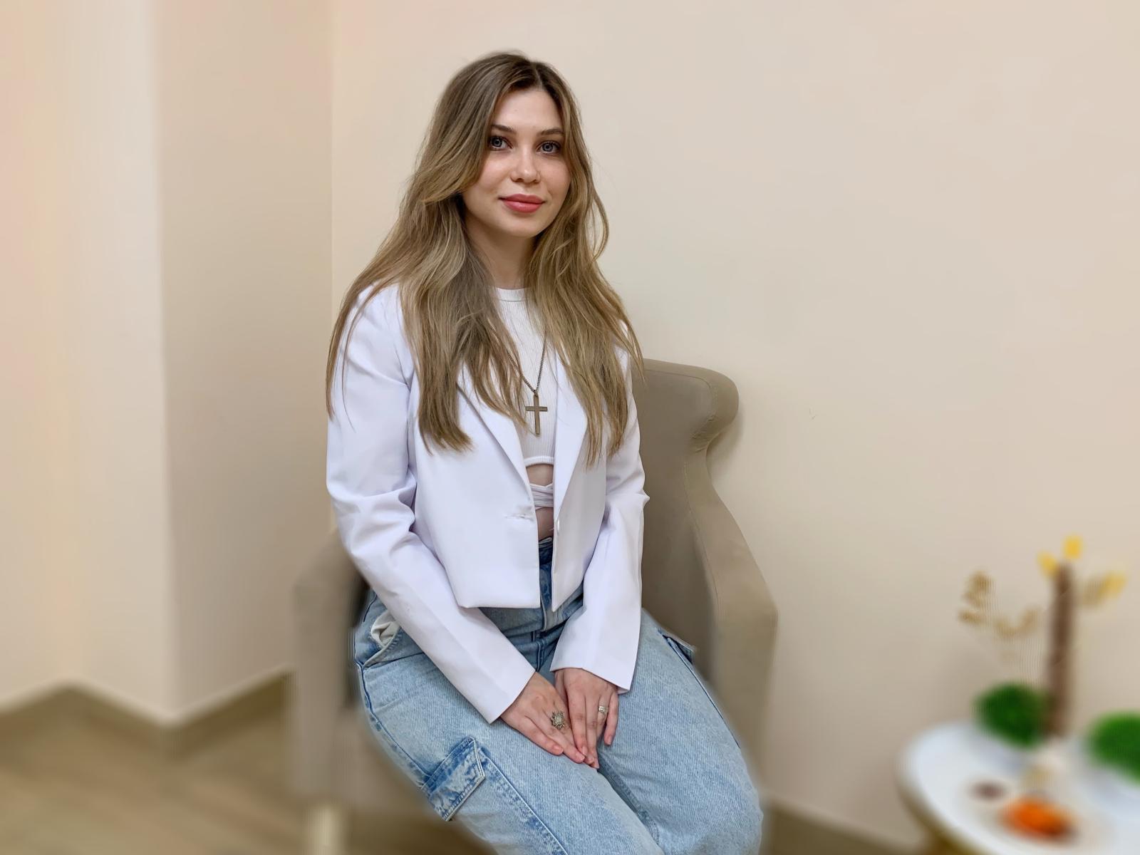 CarmingVibe with long blonde hair, white jacket, and blue jeans, sitting on a gray chair, smiling softly, hands on knees, small cross necklace, subtle makeup, relaxed vibe.