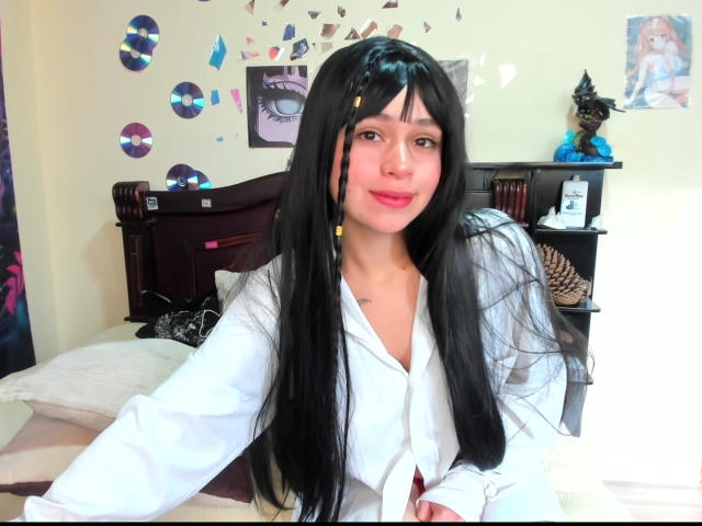 Hesttia with long black hair, wearing a white shirt, sitting on a bed, smiling, with small yellow hairpins, and a cute black cat statue in the background.