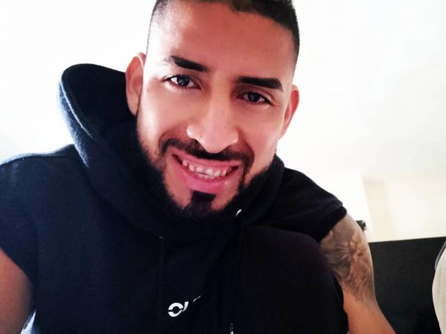 AlexSexyGamer has a cute, smiling face with dark eyebrows, a trimmed beard, and a black hoodie. He's looking at the camera with a slight tilt. Tattoo on right arm.