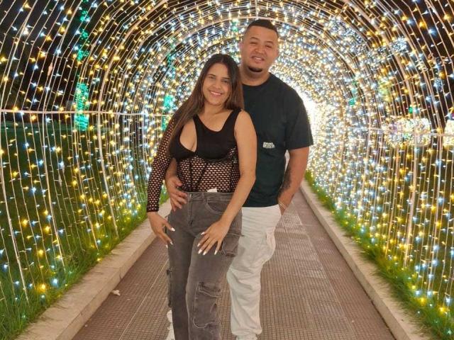 DacotaAndGrey69 with tits showing in black mesh top, brown pants, standing next to Grey69 in black shirt, white pants, arms around each other, under string lights.