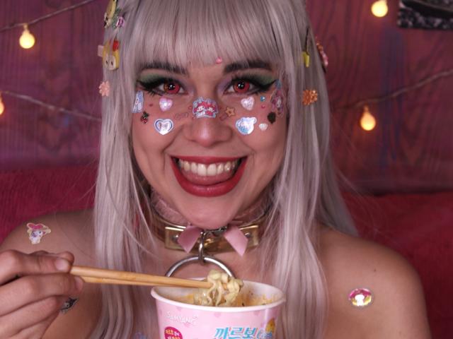 Toukauwu with blonde hair, red eyes, and heart stickers on face, smiling with tongue out, eating ramen with chopsticks, wearing pink collar with heart, in pink lighting.