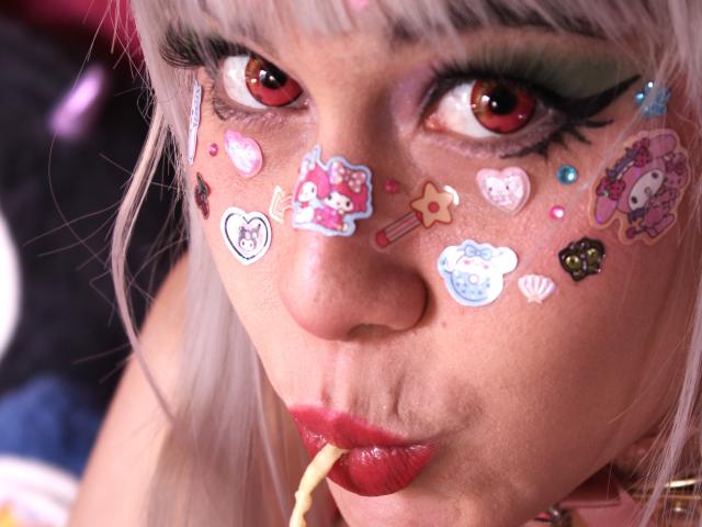 Toukauwu with pale skin, red eyes, and pink hair, sucking on a yellow lollipop. Face covered in colorful stickers: Hello Kitty, hearts, star, and more. Pouty red lips.