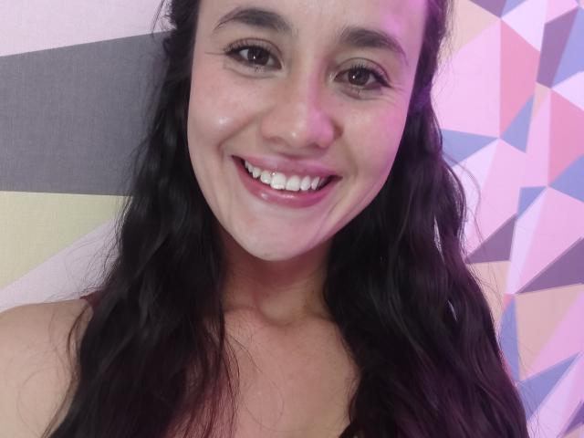 Lila69Hot has shiny skin, dark wavy hair, and a big smile showing white teeth. She's looking at the camera with pinkish-purple lighting on her face.