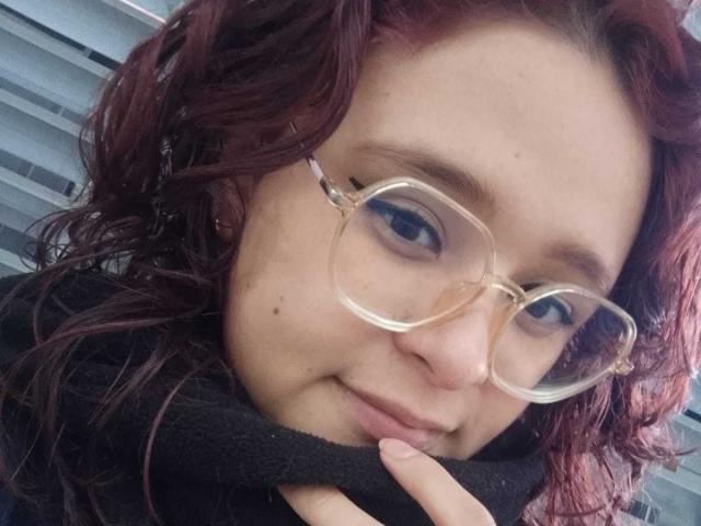 AlanisSweet has curly red hair, wears clear glasses, and touches her black scarf with a finger. She has a small mole on her right cheek and a gentle smile.