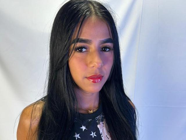 EmilyLuzz has long black hair, dark eyebrows, red lipstick, and a black star-patterned shirt. She's looking at the camera with a slight smile.
