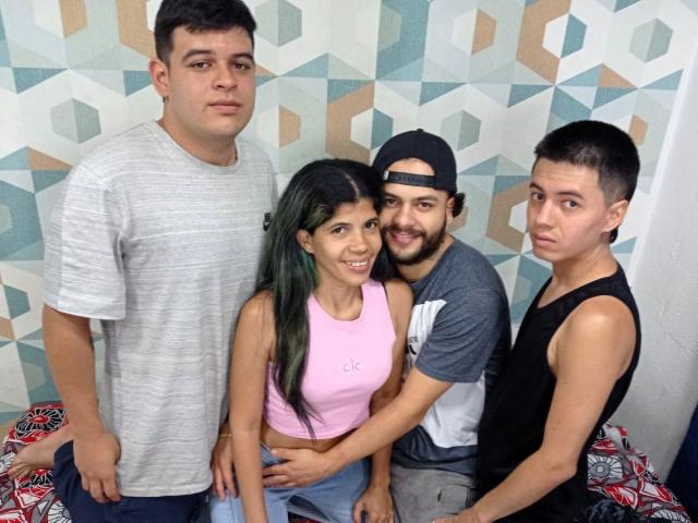 MarcelaAndBoysSexyGroup: four teenagers, standing and sitting, Marcela with green-tipped hair in pink top, boys in grey, black, and black tank tops, geometric wall pattern, casual clothes.