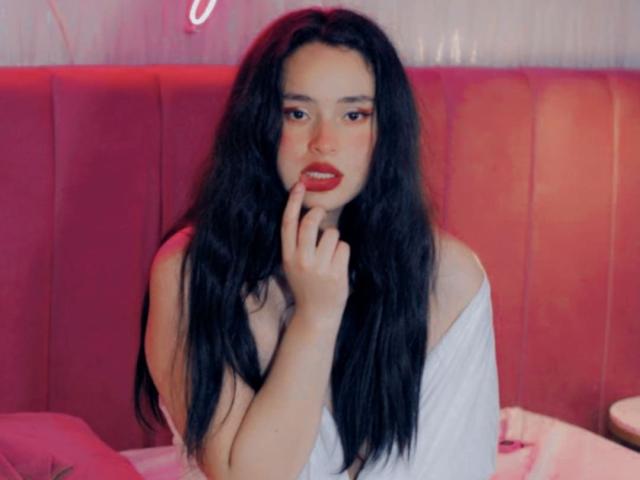 EimmyBurnys with long black hair, red lipstick, and white off-shoulder top, touching her red-painted lips with her finger, sitting in a pink, neon-lit, retro-style booth.