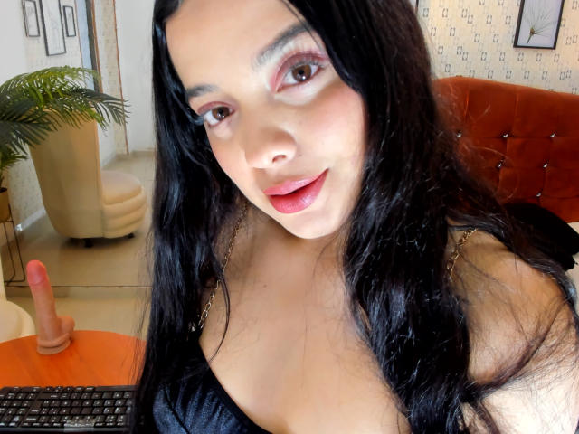 AnitaMarie69 with long black hair, pink eyeshadow, red lipstick, smiling, showing cleavage in a black top, orange couch in background, dildo on orange table.