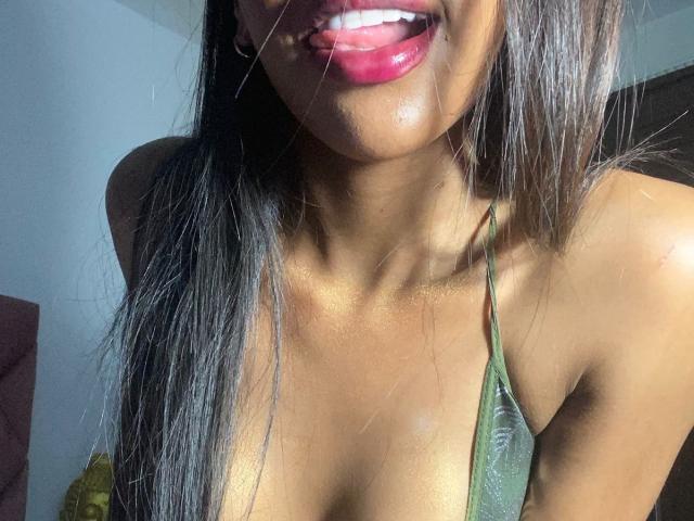 LorensX with shiny brown skin, glossy pink lips, and long black hair, wearing a green halter top, has a string of saliva from her tongue to her chin.