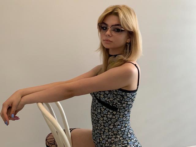 ZoeCharming with blonde hair, glasses, and a floral dress, sitting on a white chair, arms resting on the top, showing her tits and ass. Her nails are painted blue.