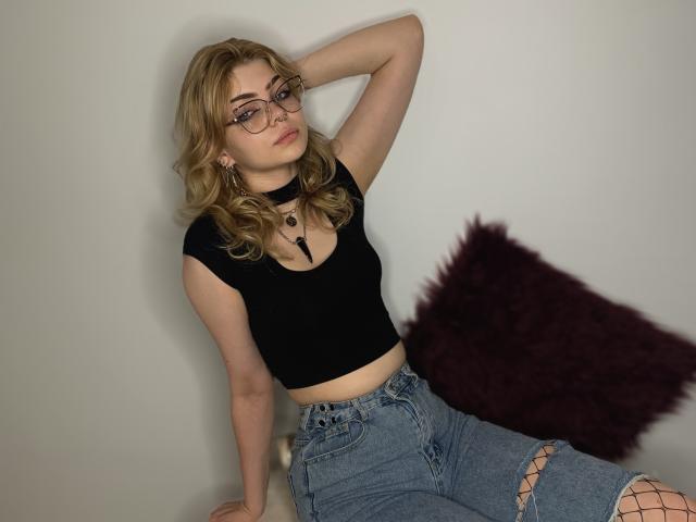 ZoeCharming with blonde hair, glasses, black crop top, and blue ripped jeans, sitting with one arm behind her head, wearing a black choker. Purple pillow on her right.