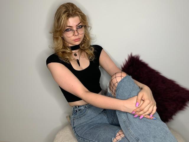 ZoeCharming with blonde wavy hair, glasses, black crop top, blue jeans, fishnet stockings, pink nails, sitting, arms around knees, dark purple pillow in background.