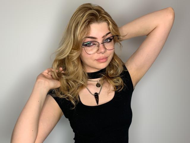 ZoeCharming has blonde wavy hair, glasses, and is wearing a black top, with her arms raised, showing her tits, and a necklace with a black pendant.