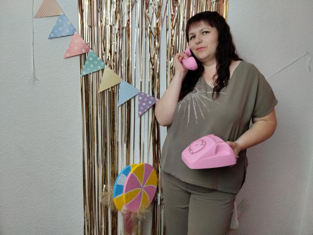 AliceInks with dark hair, green shirt, and pants, holding pink phone and pink receiver, standing, talking on phone, in front of gold string and colorful bunting.