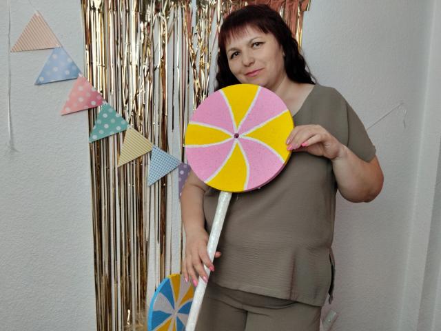 AliceInks holding a big pink, yellow, and white swirl boomerang, wearing a gray top and pants, with brown hair, standing in front of gold foil and bunting.