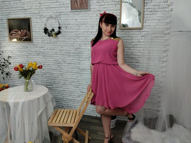 NinaFlawlees with pink dress, holding skirt, showing her ass, black hair, red hairclip, wooden chair, white brick wall, flowers, mirror, and wreath in a cozy room.
