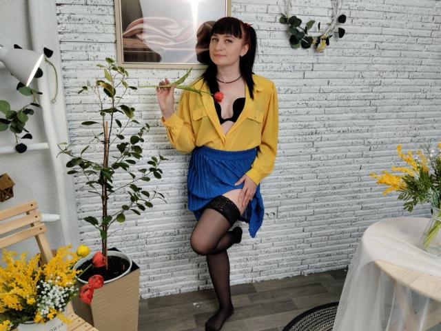 NinaFlawlees met een zwarte bh, een gele jas, een blauwe rok, zwarte kousen, die een roos vasthoudt, staand in een kamer met planten, een houten stoel en een witte bakstenen muur. Borsten zichtbaar onder de jas..