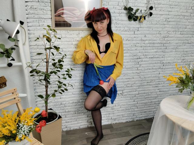 NinaFlawlees with red pigtails, yellow shirt open showing black bra, blue skirt, black thigh-highs, holding pink butt plug, standing in room with plants and yellow flowers.