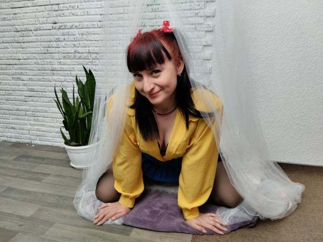 NinaFlawlees with red and black pigtails, wearing a yellow shirt, blue skirt, and sheer veil, kneeling on a purple pillow, smiling, with a plant in the background.
