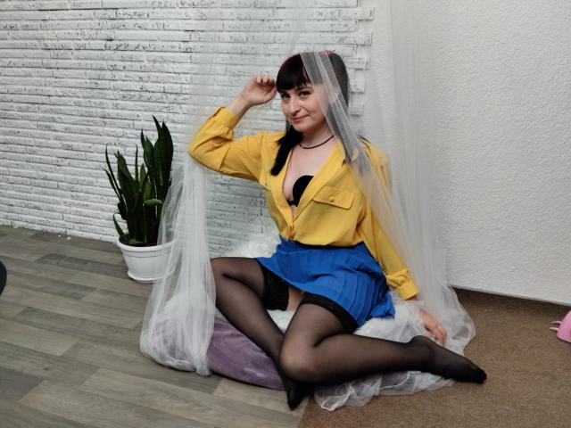 NinaFlawlees con un sujetador negro, una camisa amarilla, una falda azul, medias negras y un velo transparente, sentada sobre un cojín morado, con las piernas separadas, sonriendo y con una mano tocándose la cabeza. Sus pechos son visibles..