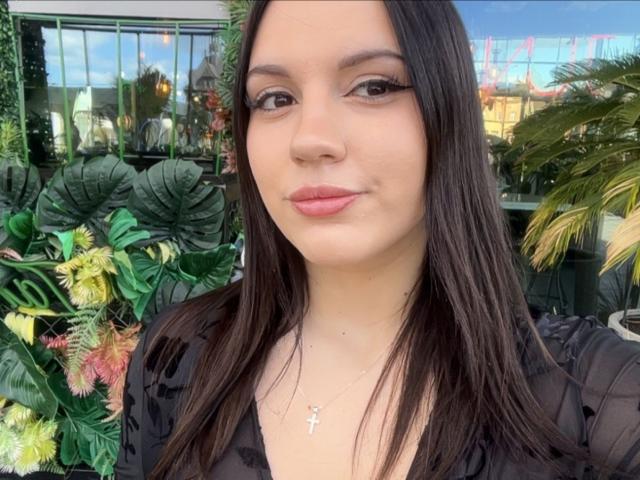 LaylaBusty69 has long, straight brown hair, light skin, and is wearing a black sheer top with a cross necklace. Her pink lips are slightly puckered in a subtle smile.