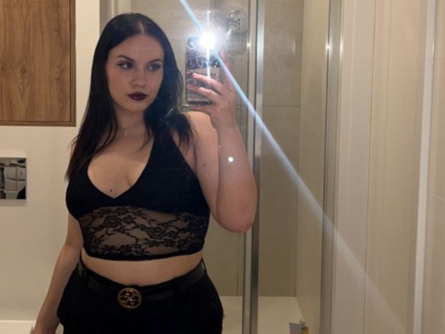 LaylaBusty69 with dark red lipstick, black lace top, and high-waisted black pants, holding phone, mirror reflection, flash, medium tits, fair skin, long black hair, serious expression.