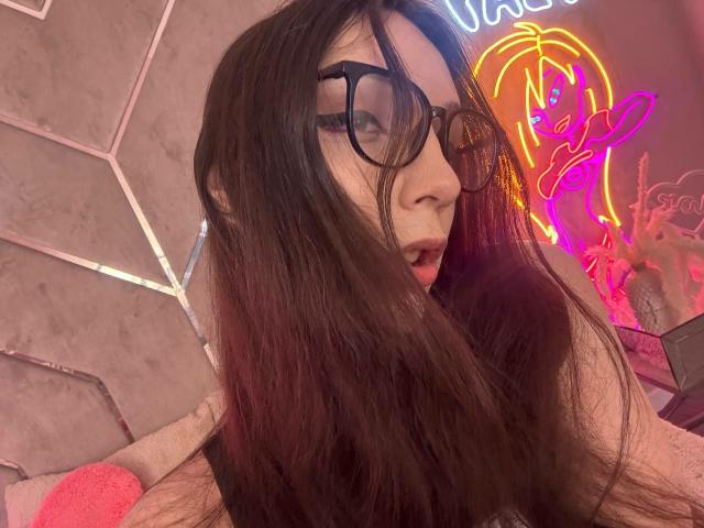 PixelShade has long brown hair, black glasses, and an open mouth with a slightly surprised expression. Neon pink and yellow light in the background. Boobs not visible.