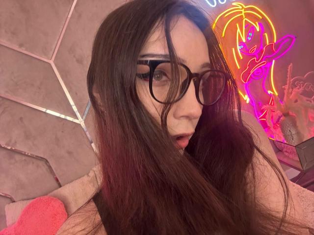 PixelShade with long brown hair, wearing black glasses, looking surprised, mouth slightly open, neon pink and yellow face outline in background, gray wall with pink geometric patterns.
