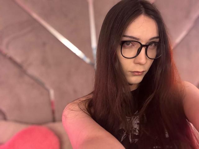PixelShade has long brown hair, black glasses, and a serious expression, showing off her tits slightly with a pink pillow in the blurry background.