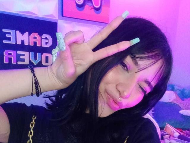 LindaWatsong with black hair, light skin, and green nails, winking, making a peace sign, and smiling with a nose ring, wearing a black shirt with gold chain.