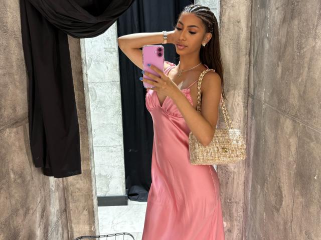 MarissaDevone with long braids, pink satin dress, holding pink phone, wearing gold bracelet, and beige woven bag, taking a selfie in a textured, brown-walled, black-draped space.