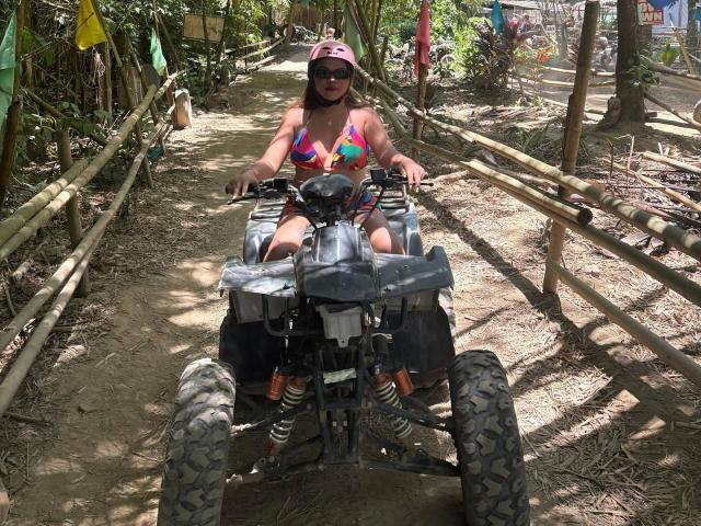 YourBestGirlX riding a black ATV with big tires, wearing a pink helmet, sunglasses, and a colorful bikini, on a dirt path with bamboo fences on both sides.