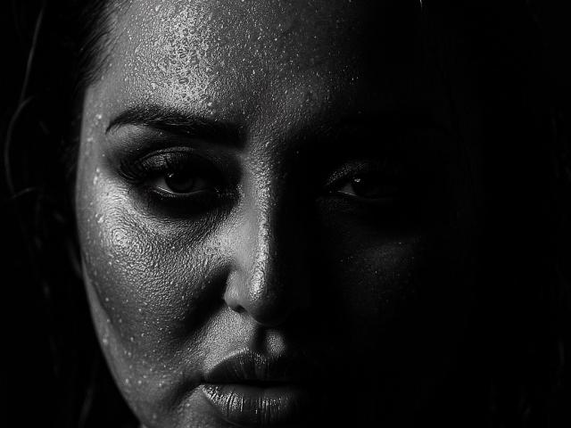 GoddessXDeborah's face in a black-and-white shot, her skin glistening with sweat, dark shadows over her right eye, and intense, focused eyes looking straight at the camera.