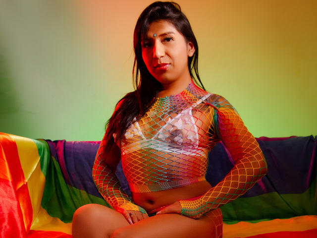 IsabellaStonnee with tan skin and long black hair, wearing a colorful fishnet top, exposing her small tits and a transparent bra, sitting on a rainbow-covered couch.